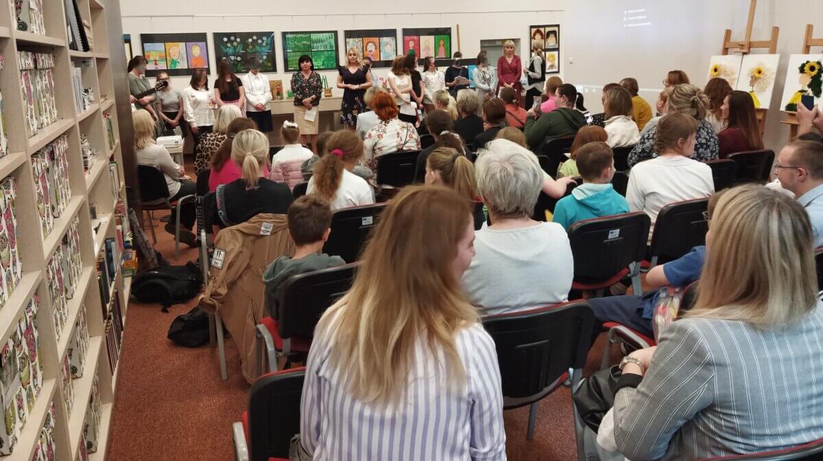 Wnętrze biblioteki, publiczność siedzi na krzesłach tyłem do obiektywu, z przodu kobieta przemawia, za jej plecami wystawa.