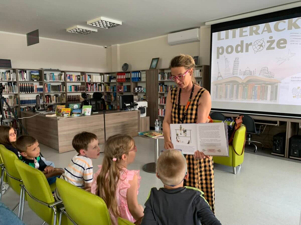 Autorka pokazuje dzieciom swoją książkę. Dzieci przygladają się z zaciekawieniem. W tle regały książkowe. Na ekranie wyświetlany jest tytuł spotkania Literackie podróze.