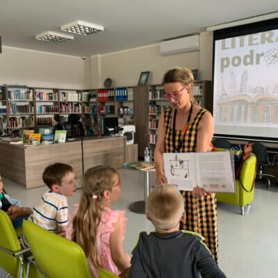 Autorka pokazuje dzieciom swoją książkę. Dzieci przygladają się z zaciekawieniem. W tle regały książkowe. Na ekranie wyświetlany jest tytuł spotkania Literackie podróze.