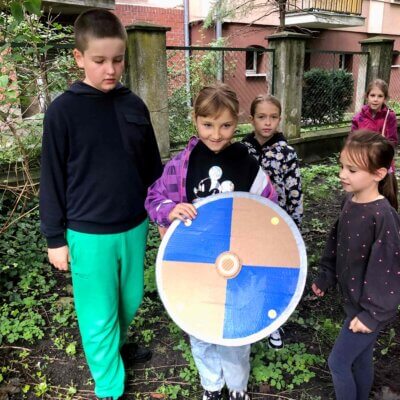 Dzieci w parku stoją i prezentują znalezioną antyczną tarczę. Kliknięcie powoduje powiększenie zdjęcia