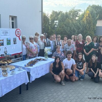 Grupa uczestników na dziedzińcu biblioteki przy tablicy wystawienniczej dotyczącej książki „Nad Niemnem", stolikach z książkami i poczęstunkiem. Klikniecie powoduje powiększenie zdjęcia.