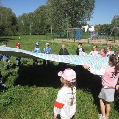 Dziewiętnaścioro dzieci w wieku 5 lat, stojące na trawie obok placu zabaw, trzymające wielki piknikowy, turkusowy koc we flamingi. Kliknięcie w obrazek spowoduje powiększenie.