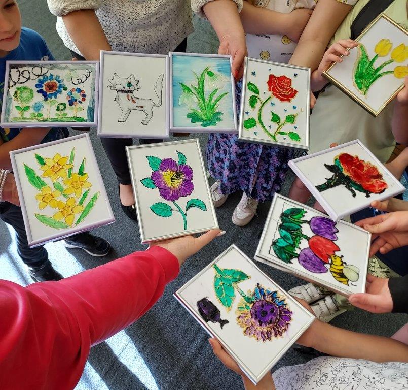 Uczestnicy warsztatów malowania na szkle prezentują wykonane przez siebie prace przedstawiające różnorodne, kolorowe kwiaty oraz kota.