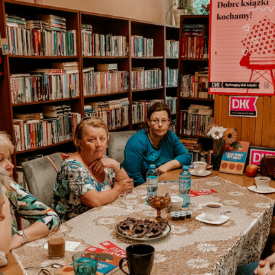 Wnętrze biblioteki. Trwa spotkanie Dyskusyjnego Klubu Książki. Przy stole siedzi sześć kobiet. Na stole leży omawiana książka „Z nieba spadły trzy jabłka" Narine Abgarjan. W tle plakat promujący DKK. Kliknięcie powoduje powiększenie zdjęcia.