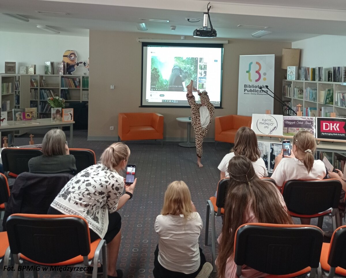 Dziewczynka w ekwilibrystycznej pozycji wśród widzów w pomieszczeniu biblioteki.