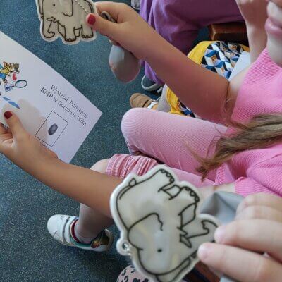 Dzieci siedzą i trzymają w ręce odblaskowe słonie i karteczki z odbiciem swoich linii papilarnych. Kliknięcie powoduje powiększenie zdjęcia.