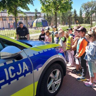 Auto policyjne, na które patrzy grupa dzieci. Za autem stoi pani policjantka. Kliknięcie powoduje powiększenie zdjęcia.