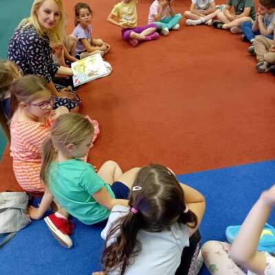 Dzieci siedzą na dywanie, a bibliotekarka czyta im książkę. Kliknięcie powoduje powiększenie zdjęcia.