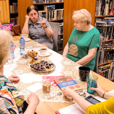 Dyskusyjny Klub Książki w Filii nr 8. Sześć czytelniczek siedzi w bibliotece przy stole. Jedna z kobiet gestykuluje, pozostałe patrzą na nią. Kliknięcie powoduje powiększenie zdjęcia.