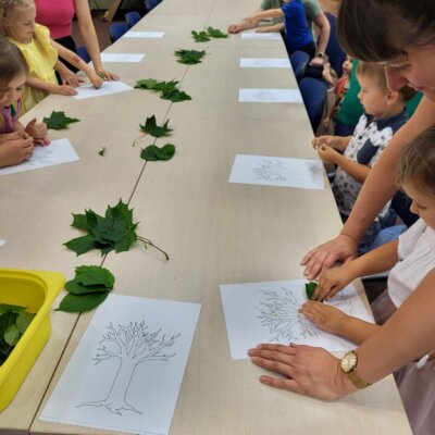Dzieci przy pomocy rodziców kolorują kartkę liśćmi. Klikniecie powoduje powiększenie zdjęcia.