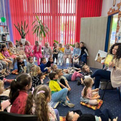 Duża grupa dzieci słucha bajki czytanej przez kobietę. Kliknięcie powoduje powiększenie zdjęcia.