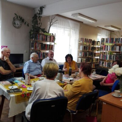 członkowie Dyskusyjnego Klubu Książki siedzą przy stole z poczęstunkiem i książkami, gadżetami DKK, w tle kwiaty. Kliknięcie w obrazek spowoduje powiększenie.