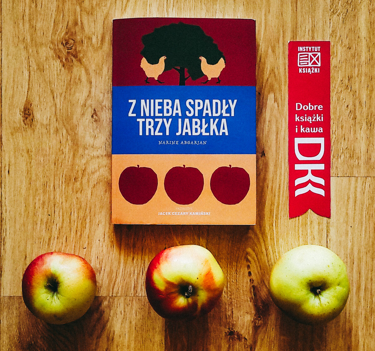 W centrum leży książka „Z nieba spadły trzy jabłka" Narine Abgarjan. Po prawej różowa zakładka do książki z napisem Dobre książki i kawa. Na dole trzy jabłka nawiązujące do okładki książki