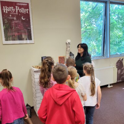 Bibliotekarka wskazuje dzieciom regał z literaturą o Harrym Potterze. Kliknięcie powoduje powiększenie zdjęcia.