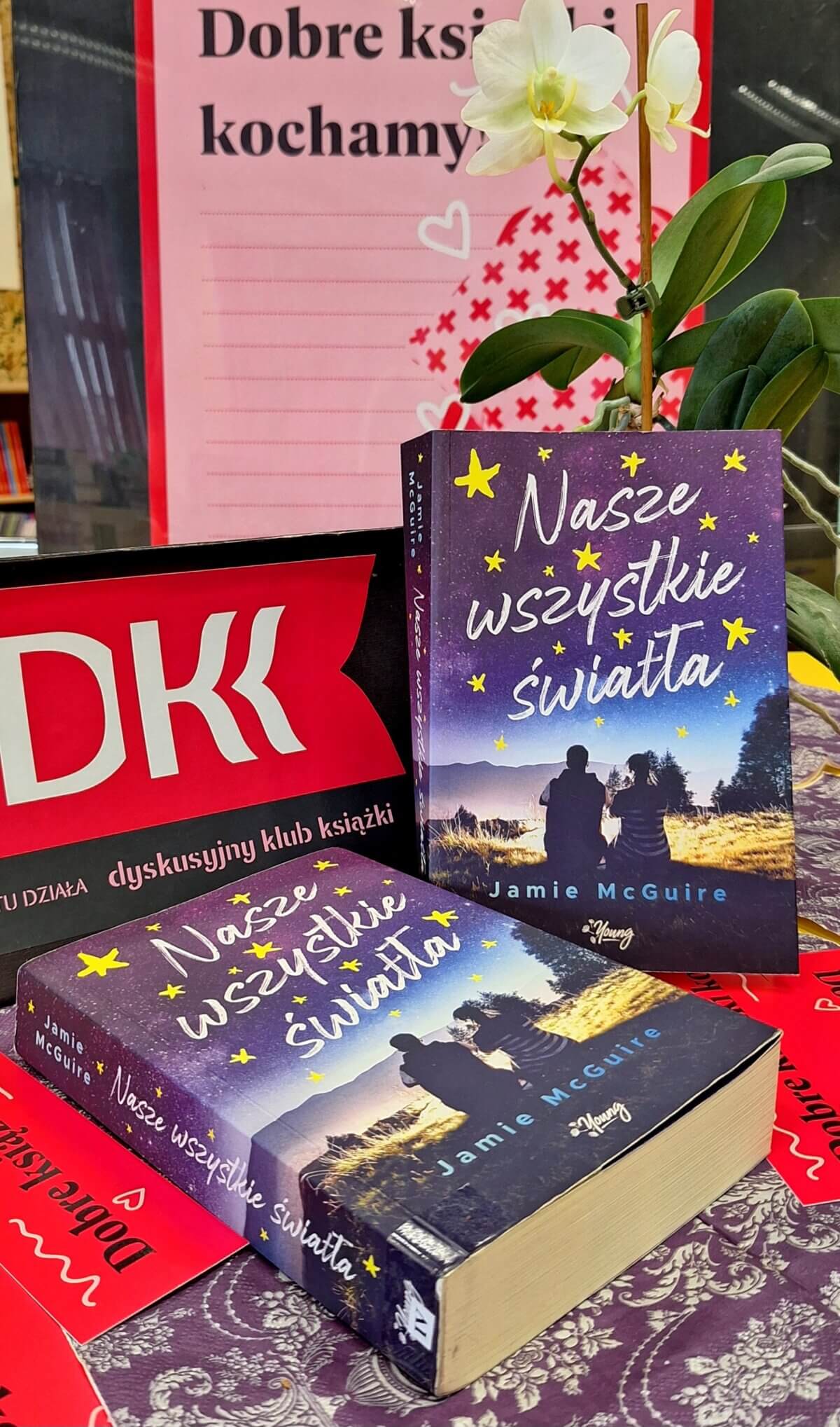 Na stole leżą dwie książki „Nasze wszystkie światła”, za nimi stoi storczyk, a w tle plakat DKK.