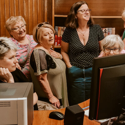 Siedem kobiet patrzy w monitor komputera położony na biurku. Niektóre z nich się uśmiechają. Widać zaciekawienie na ich twarzach. Kliknięcie powoduje powiększenie zdjęcia.