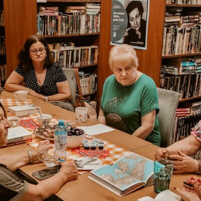 Biblioteka. Trwa spotkanie Dyskusyjnego Klubu Książki. Siedem kobiet rozmawia na temat omawianej pozycji. Na stole leżą ciastka, zakładki i kubki. Kliknięcie powoduje powiększenie zdjęcia.