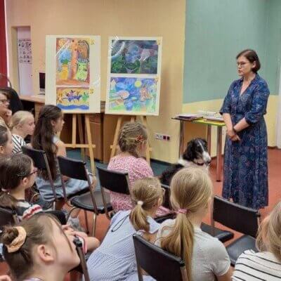 Autorka Anna Czerwińska-Rydel stoi. Przed nią siedzą dzieci. Po lewej stronie są nagrodzone prace na sztalugach. Kliknięcie powoduje powiększenie zdjęcia.