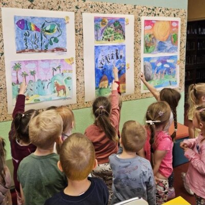 Przedszkolaki stoją tyłem do wystawy. Pokazują kolorowe prace. Kliknięcie w obrazek spowoduje powiększenie.