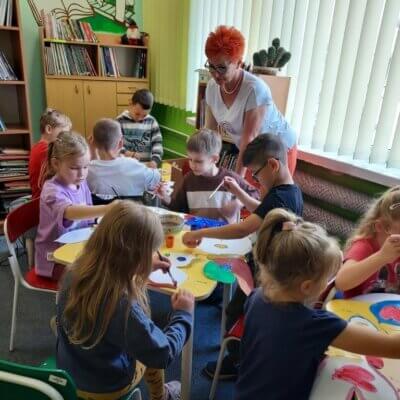 Dzieci siedzące przy stolikach malują farbami wycięte z papieru motyle i pomagająca im w pracy Bożena Ambroży. Kliknięcie w obrazek spowoduje powiększenie.
