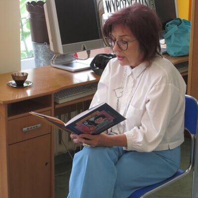 Kobieta siedząca na niebieskim krześle czytająca książkę w tle biurko z monitorem. Kliknięcie powoduje powiększenie zdjęcia.