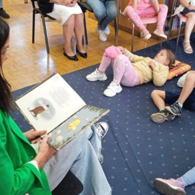Kobieta siedzi na krześle i czyta bajkę dzieciom siedzącym na krzesłach i na dywanie. Kliknięcie w obrazek spowoduje powiększenie.