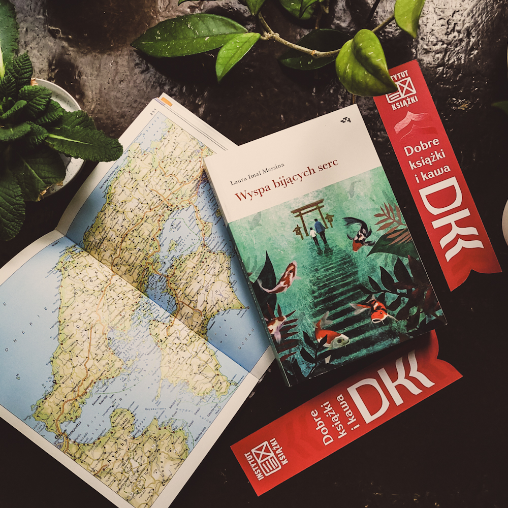 Na ciemnym tle dwie różowe zakładki DKK, książka otworzona na otwartej mapie Japonii oraz „Wyspa bijących serc” L. I. Messina, czyli omawiana pozycja.
