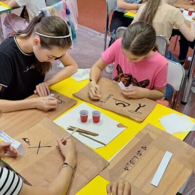 Dzieci siedzą przy stole i na przygotowanych przez siebie papirusach piszą hieroglify tuszem i patykiem. Kliknięcie powoduje powiększenie zdjęcia.