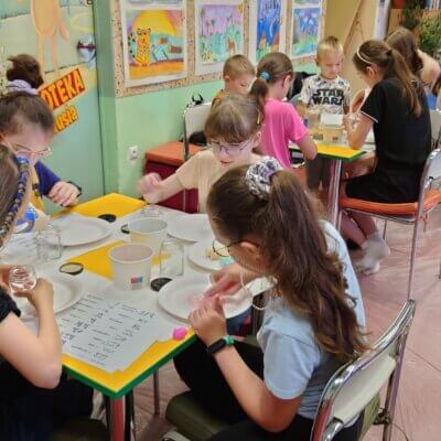 Dzieci siedzą przy stole i barwią sól kolorową kredą. Kliknięcie powoduje powiększenie zdjęcia.