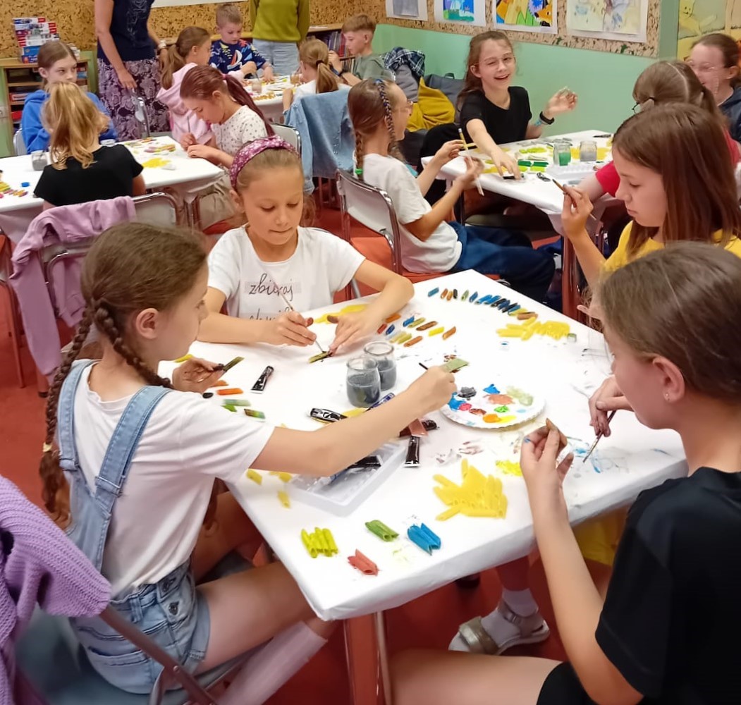 Dzieci siedzą przy stolikach i malują kolorowymi farbami makaron.