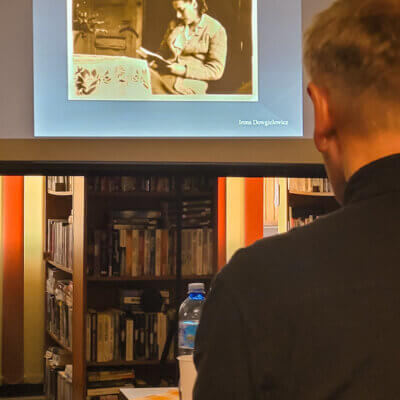 Wnętrze biblioteki. Widok na rzutnik i slajd wyświetlanej prezentacji. To zdjęcie Ireny Dowgielewicz czytającej książkę przy stole. Na pierwszym planie prowadzący spotkanie stoi tyłem. Kliknięcie w obrazek spowoduje powiększenie.