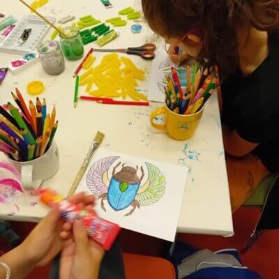 Na stole leżą kredki, makaron i pokolorowane skarabeusze. Dziewczynka maluje swojego żuka. Kliknięcie powoduje powiększenie zdjęcia.