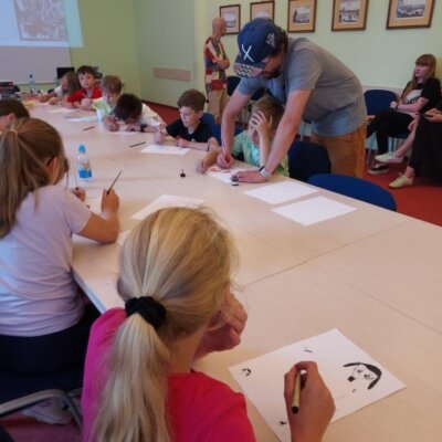 Jacek Fraś pomaga dzieciom siedzącym przy stole w rysowaniu postaci z mangi. Kliknięcie powoduje powiększenie zdjęcia.