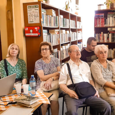 Wnętrze biblioteki. Po lewej prowadzący spotkanie Janusz Dreczka, na wprost zebrana publiczność. Na krzesłach siedzi sześć osób. Na stole kwiaty, laptop. Kliknięcie w obrazek spowoduje powiększenie.