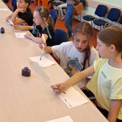 Dziewczynki siedzą przy stole i piszą gęsim piórem oraz atramentem. Kliknięcie powoduje powiększenie zdjęcia.