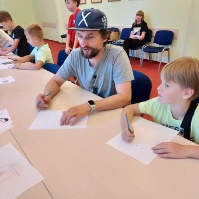 Jacek Fraś siedzący z dziećmi przy stole, na którym leżą kartki do rysowania. Kliknięcie powoduje powiększenie zdjęcia.