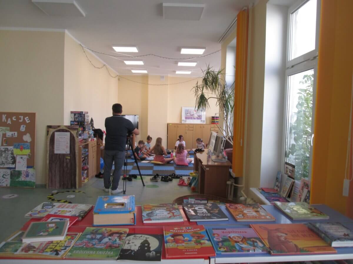 wnętrze biblioteki dla dzieci, na dalszym planie jest grupa dzieci siedząca na macie w niebiesko-czarną kratę; przed nimi widać sylwetkę kamerzysty od tyłu; na pierwszym planie na regale ułożone są książki dla dzieci.