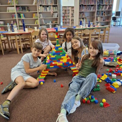 Zdjęcie przedstawia pięcioro dzieci siedzących na podłodze w bibliotece. Dzieci zbudowały dużą konstrukcję z kolorowych klocków, która przypomina wieżę Eiffla. W tle widać regały z książkami oraz stoły i krzesła. Kliknięcie powoduje powiększenie zdjęcia.