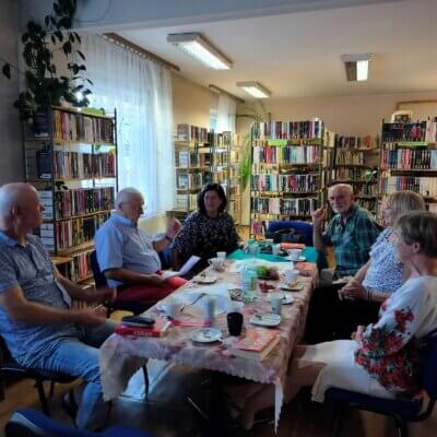 uczestnicy Dyskusyjnego Klubu Książki siedzą przy stole z poczęstunkiem i egzemplarzami książki Andrew Ridkera „Altruiści. W tle gadżety DKK. Kliknięcie powoduje powiększenie zdjęcia.