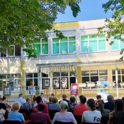 W bibliotecznym ogrodzie siedzi publiczność na krzesłach, tyłem do obiektywu. Przed nią budynek i sztalugi. Kliknięcie powoduje powiększenie zdjęcia.