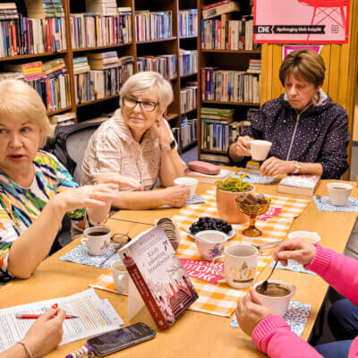 Biblioteka. Filia nr 8. Trwa spotkanie Dyskusyjnego Klubu Książki. Przy stole siedzi siedem kobiet. Na stole leży omawiana pozycja: „Kiedy zakwitną brzoskwinie" Melissy Fu. Kobiety zwrócone w kierunku moderatorki spotkania. Jedna z kobiet gestykuluje. Kliknięcie powoduje powiększenie zdjęcia.