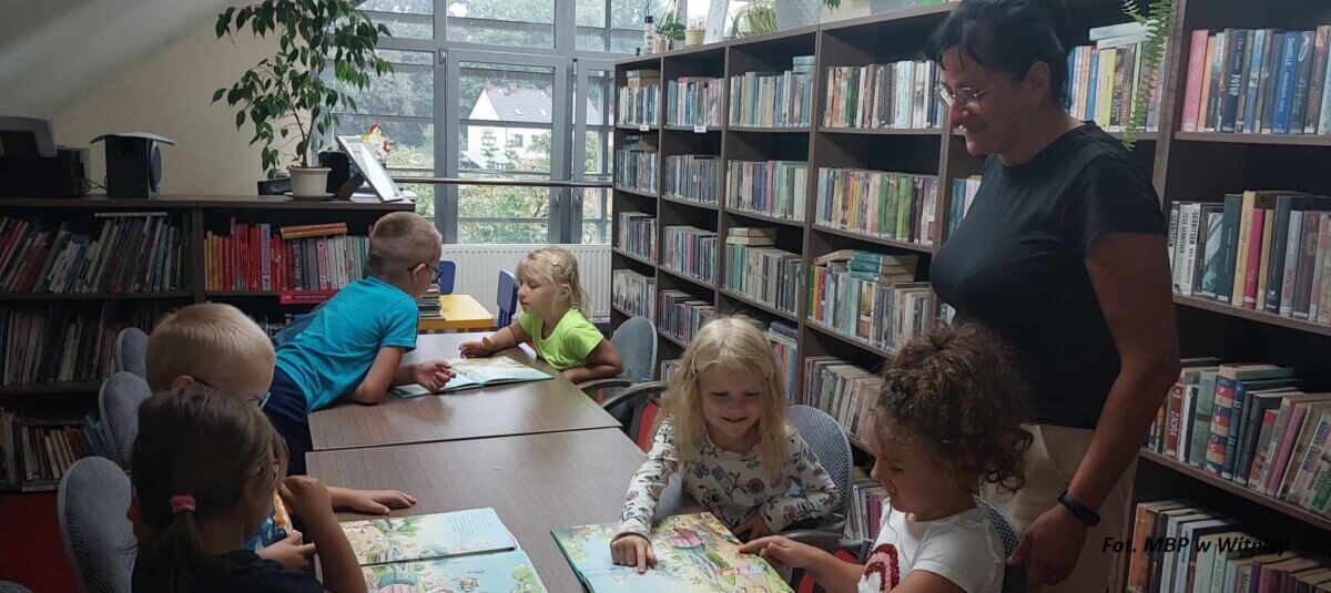 Dzieci siedzące przy stole wraz z bibliotekarką w pomieszczeniu biblioteki.