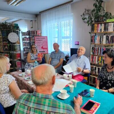 Inne ujęcie, wnętrze biblioteki, w otoczeniu stołu, przy którym siedzą uczestnicy spotkania, regały z książkami, uczestnicy Dyskusyjnego Klubu Książki siedzą przy stole z poczęstunkiem i egzemplarzami książki Andrew Ridkera „Altruiści. W tle gadżety DKK. Kliknięcie powoduje powiększenie zdjęcia.