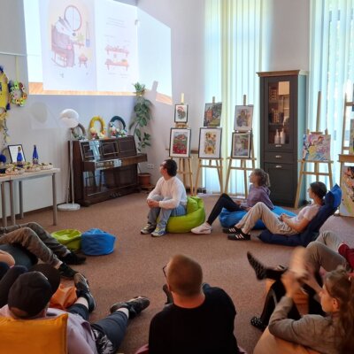 Wnętrze biblioteki, dzieci siedzą na wykładzinie na pufach i oglądają wyświetlaną na ścianie prezentację. Bibliotekarka siedzi z prawej stronie przed komputerem. Kliknięcie powoduje powiększenie zdjęcia.