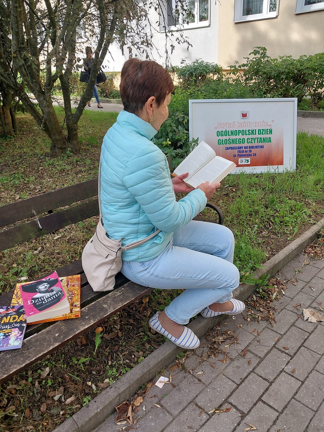 Plener, brązowa ławka na niej książki i kobieta czytająca książkę. W tle baner promujący Ogólnopolski Dzień Głośnego Czytania, ściana bloku i rośliny.