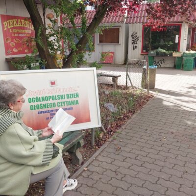 Plener, zielona ławka, kobieta czytająca książkę. W tle baner promujący Ogólnopolski Dzień Głośnego Czytania, warzywniak i rośliny. Kliknięcie powoduje powiększenie zdjęcia.