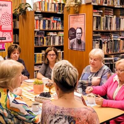 Wnętrze biblioteki. Kobiety siedzą przy stole i dyskutują na temat omawianej pozycji. W tle regały z książkami. Kliknięcie powoduje powiększenie zdjęcia.