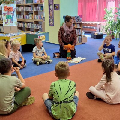 Dzieci i dorośli siedzą w kółku na podłodze. Bibliotekarka trzyma kukiełkę Plastusia. Chłopiec obok opowiada. Kliknięcie powoduje powiększenie zdjęcia.