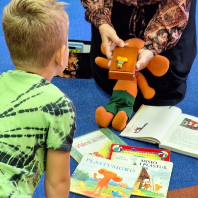 Na podłodze leżą książki o Plastusiu. Bibliotekarka trzyma w ręku drewniany piórnik. Na nim leży plastelinowy ludzik. Kliknięcie powoduje powiększenie zdjęcia.