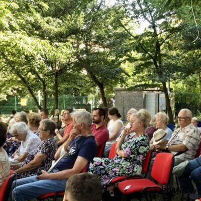 Goście spotkania siedzą na czerwonych krzesłach w plenerze. W tle liczne zielone drzewa. Kliknięcie powoduje powiększenie zdjęcia.